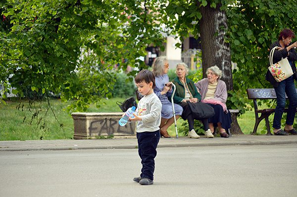 boy holding bottle in front of older women sitting on park bench