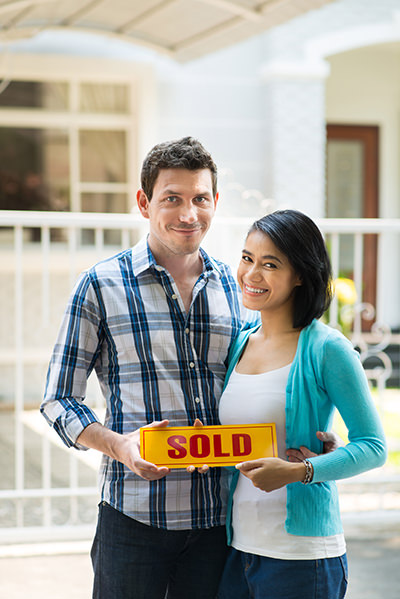 picture of a heterosexual couple standing in front of a house holding a sign that says Sold