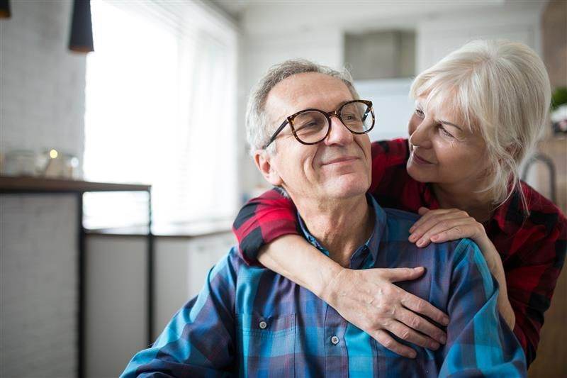 An elderly couple embrace in a living room, smiling at each other