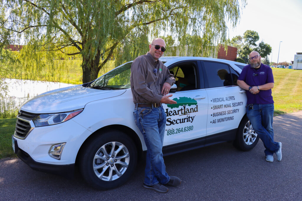Two men stand next to a white car with the Heartland Security logo on it