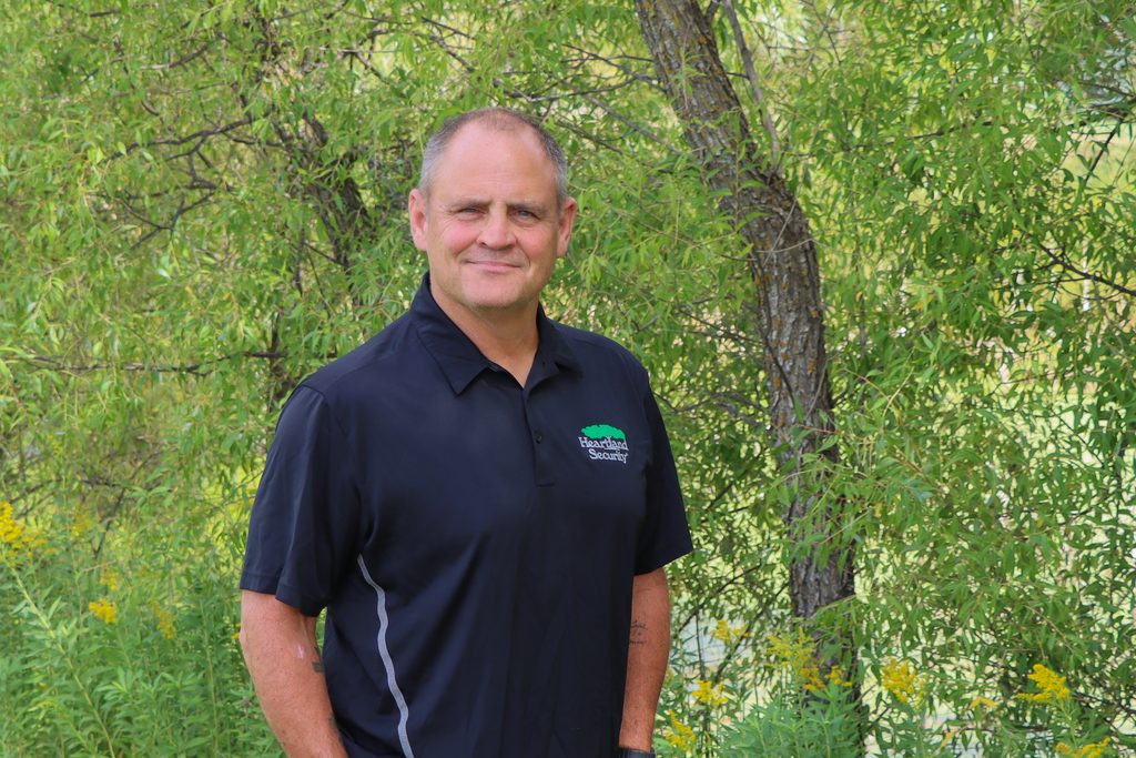 A man in a black shirt with the Heartland logo on it stands in front of a green nature scene